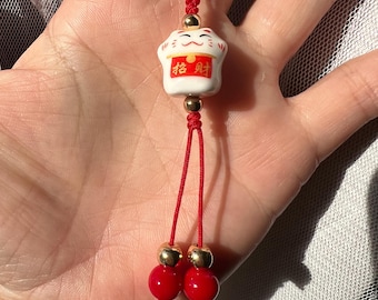 May include: A red string charm with a white ceramic cat figurine holding a red envelope with gold Chinese characters that read "good fortune". The charm has two red beads with gold accents.