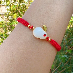 May include: A red string bracelet with a white jade rabbit charm. The rabbit has gold ears and a red eye.