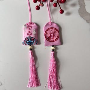 May include: Two pink fabric charms with tassels. One charm features a colourful floral design and text, the other has a red circular design and text. Both charms have pink cords and tassels. The text "Pink-Love/Relationship" is at the bottom.