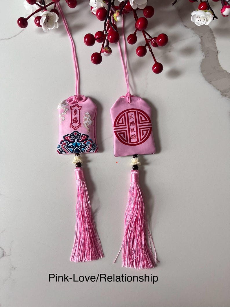 May include: Two pink fabric charms with tassels. One charm features a colourful floral design and text, the other has a red circular design and text. Both charms have pink cords and tassels. The text "Pink-Love/Relationship" is at the bottom.