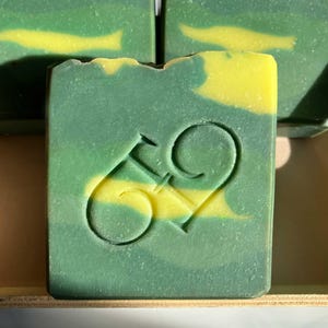 May include: A square bar of handmade soap with a green and yellow swirl design. The number "69" is carved into the front of the soap. The soap is part of a collection of similar bars.