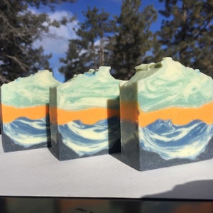 May include: Three bars of handmade soap with a layered design. The top layer is a light green color, the middle layer is a bright orange color, and the bottom layer is a dark blue and white swirl.