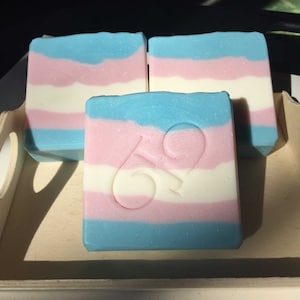 May include: Three square soaps with a blue, pink, and white striped design, resembling a flag. One soap has the number "69" carved into it. The soaps are arranged on a light wooden tray.