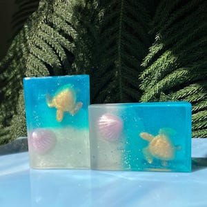 May include: Two clear blue and white soap bars with a glittery finish. Each bar features a golden turtle and a pink seashell.