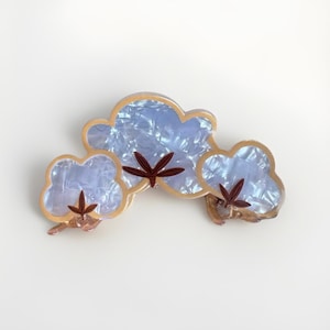 Op de afbeelding: Een blauwe en gouden haarspeld met een wolkendesign. De wolken hebben een parelmoer afwerking en zijn versierd met bruine katoenen bloemdetails.