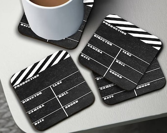 May include: Four square coasters with a black clapperboard design. Each coaster has white text including "PRODUCTION", "DIRECTOR", "CAMERA", "DATE", "TAKE", "ROLL", and "SOUND". A white mug sits on one coaster.