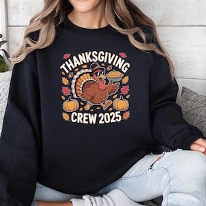 May include: Black sweatshirt with a Thanksgiving-themed graphic. The design features a cartoon turkey wearing sunglasses and a hat, holding a pie. The text reads "Thanksgiving Crew 2025" with pumpkins and fall leaves.
