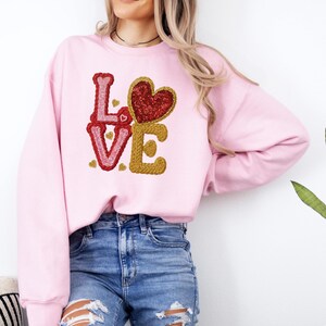 May include: A light pink sweatshirt featuring the word "LOVE" in a textured, embroidered design. The "L" and "V" are in red, the "O" is a red sequined heart, and the "E" is in gold. The sweatshirt is a casual style.