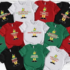 May include: A collection of short-sleeved t-shirts in various colors, including black, white, red, and green. Each shirt features a graphic of an elf with text. The text includes phrases like "He must be a South Pole Elf" and "Somebody Hug".
