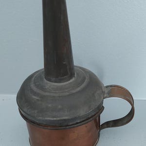 Puede incluir: Lámpara de aceite antigua de cobre con boquilla de metal oscuro y asa. La lámpara tiene una base redonda y una boquilla cilíndrica. El asa es un simple lazo. La lámpara probablemente se utiliza para iluminación o decoración.