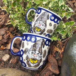Puede incluir: Dos tazas de cerámica azul y blanca con un diseño de gnomo. Cada taza presenta un gnomo sosteniendo una taza amarilla con la palabra "Coffee" en ella. Las tazas están rodeadas de follaje marrón y verde.