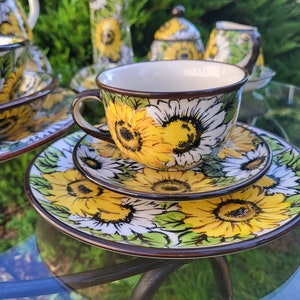 Puede incluir: Juego de té de cerámica con diseño de girasoles. El juego incluye una taza, un platillo y un plato, todos adornados con girasoles amarillos y blancos y hojas verdes. Los bordes son marrones. Incluye azucarero y jarrón.