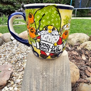 May include: A blue and white ceramic mug featuring a gnome holding a banner that reads "World's Best Mom". The gnome is wearing a green hat and red trousers. The mug is decorated with flowers and leaves.
