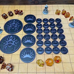 Puede incluir: Un juego de mesa con dados, figuras en miniatura y fichas azul oscuro con un diseño de pulpo. Las piezas del juego están dispuestas sobre una superficie con patrón de cuadrícula, lo que sugiere un juego de rol o estrategia.