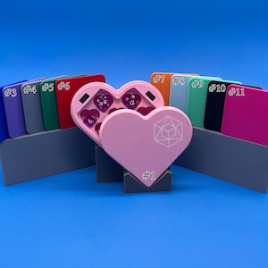 May include: A pink heart-shaped dice tower with a white geometric design. The tower is open and shows purple dice inside. The tower is surrounded by eleven small, rectangular boxes, each with a number from 1 to 11.