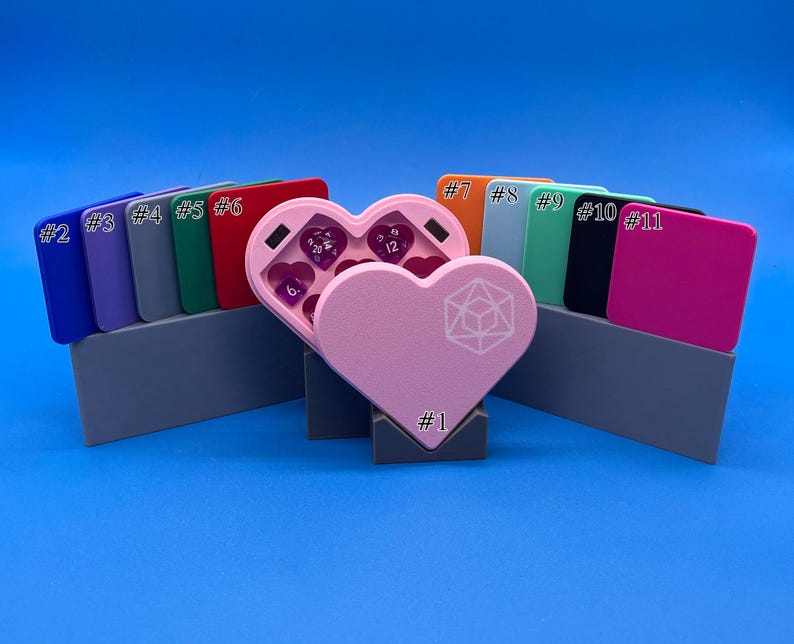 May include: A pink heart-shaped dice tower with a white geometric design. The tower is open and shows purple dice inside. The tower is surrounded by eleven small, rectangular boxes, each with a number from 1 to 11.