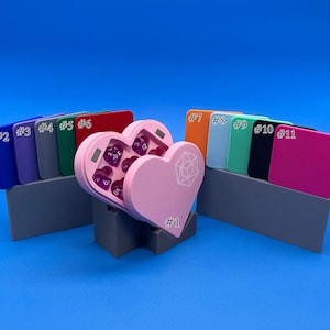 May include: A pink heart-shaped dice box with a clear lid, open to reveal purple dice. The box is on a grey stand with the number "#1" printed on it. There are 11 colour swatches behind the box, labelled with numbers 2 through 11.