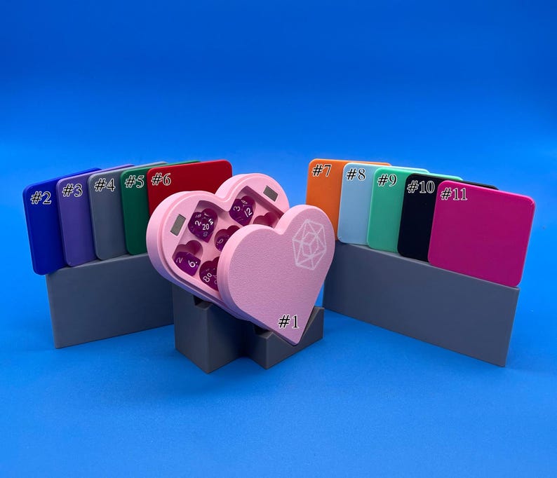 May include: A pink heart-shaped dice box with a clear lid, open to reveal purple dice. The box is on a grey stand with the number "#1" printed on it. There are 11 colour swatches behind the box, labelled with numbers 2 through 11.