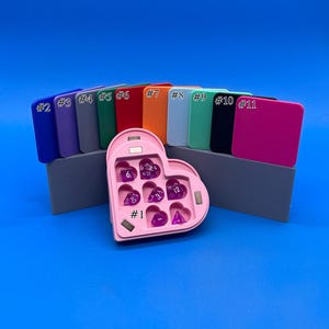 May include: A pink heart-shaped dice tray with seven purple dice inside. The tray has a magnetic closure and is numbered #1. The tray is sitting on a grey surface in front of eleven different coloured squares.