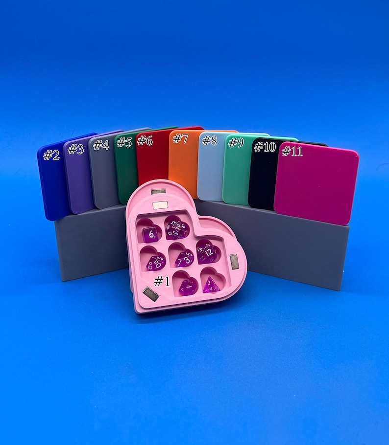 May include: A pink heart-shaped dice tray with seven purple dice inside. The tray has a magnetic closure and is numbered #1. The tray is sitting on a grey surface in front of eleven different coloured squares.