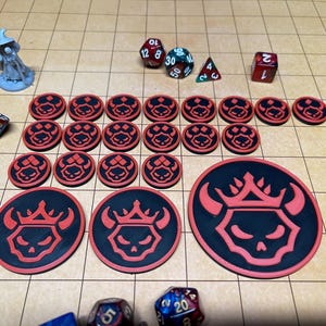 May include: A collection of tabletop gaming accessories, including dice of various shapes and colors, and red and black tokens with a skull and crown design. A miniature wizard figure is also present.