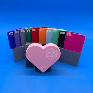 May include: A set of 11 colour swatches in various shades, including blue, purple, green, red, orange, light blue, green, black, and pink. The swatches are arranged in a semi-circle shape with a pink heart-shaped object in the centre. The heart has a white geometric design.