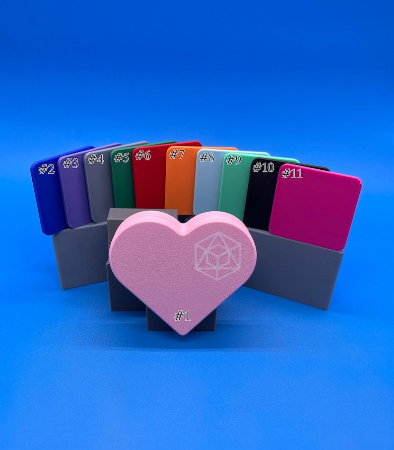 May include: A set of 11 colour swatches in various shades, including blue, purple, green, red, orange, light blue, green, black, and pink. The swatches are arranged in a semi-circle shape with a pink heart-shaped object in the centre. The heart has a white geometric design.