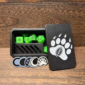 May include: A black rectangular dice box with a bear paw and leaf emblem on the lid. Inside are bright green polyhedral dice and a slotted tray. Five small circular tokens with moon, paw, and circle designs are arranged in front of the box.