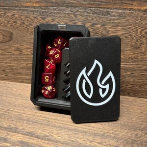 May include: A black rectangular dice box with a white flame icon on the sliding lid. The open box reveals a set of translucent red polyhedral dice with gold numbers, featuring glittery inclusions, stored in a molded black interior compartment.