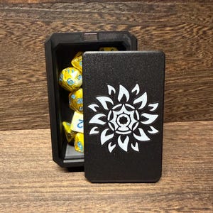 May include: A rectangular black dice box with a magnetic lid featuring a white engraved sunburst design. The open box reveals a collection of yellow polyhedral dice with blue numbers, showcasing various geometric shapes used for tabletop gaming.