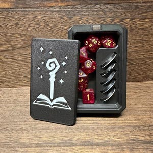 May include: A black rectangular dice box with a magnetic lid featuring a white graphic of a staff and an open book. The interior contains a set of translucent red glitter polyhedral dice with gold numbers and five black circular tokens.