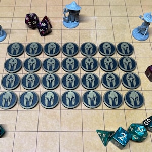May include: A tabletop gaming scene featuring blue and teal dice, miniature figures, and round tokens with a helmet design. The game pieces are arranged on a grid-patterned surface, suggesting a role-playing game.
