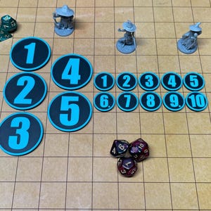 May include: A tabletop gaming setup featuring numbered tokens, miniature figurines, and various dice. The tokens are black with light blue numbers. The dice are in shades of green, red, and black. The game pieces are arranged on a grid.
