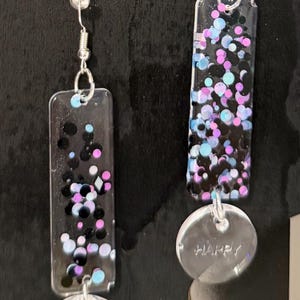 May include: A pair of dangle earrings with a rectangular top and a circular bottom. The rectangular portion is black with colorful confetti. The circular bottom has the word "HAPPY" etched on it.