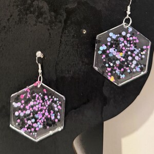 May include: Hexagon-shaped earrings with a clear resin base, filled with colorful glitter. The glitter includes pink, purple, blue, and black specks. The earrings have silver-colored hooks.