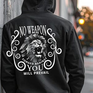Puede incluir: Sudadera con capucha negra con un gráfico de león blanco y el texto "NO WEAPON FORGED AGAINST YOU WILL PREVAIL." La sudadera tiene una capucha con cordón. La persona está apoyada en una pared gris.