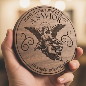 Può includere: Un sottobicchiere rotondo in legno con un'incisione di un angelo. Il testo "TODAY IN THE TOWN OF DAVID A SAVIOR HAS BEEN BORN TO YOU" è inciso sul bordo. L'angelo ha le ali spiegate.