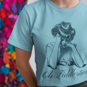 May include: A light blue t-shirt with a vintage illustration of a woman with her head in her hand. The text "Oh Fiddlesticks" is printed below the illustration.