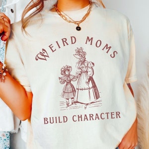 May include: Cream-coloured t-shirt with the text "WEIRD MOMS BUILD CHARACTER" in a burgundy font. The shirt features a vintage illustration of a woman and child in dresses and hats. The shirt has short sleeves and a relaxed fit.