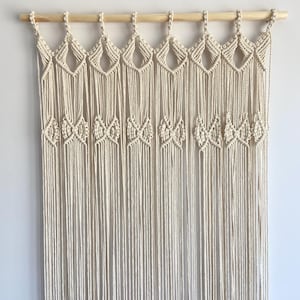 May include: A cream-coloured macrame wall hanging suspended from a light-toned wooden dowel. The design features a series of diamond-shaped knots at the top, followed by a row of smaller, intricate bow-like knot patterns, ending in long, vertical fringe.