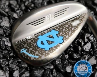 Puede incluir: Un wedge de palo de golf plateado con un logotipo azul claro y la palabra "Sweetspot". El palo tiene las letras "BV" y "SM9" grabadas. El palo está decorado con un patrón repetitivo. El palo está sobre una superficie oscura.