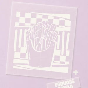 Può includere: Uno stencil quadrato semitrasparente con un grafico centrale di patatine fritte in un cartone, su uno sfondo a scacchi e righe. Uno stencil rettangolare più piccolo e separato include il testo "pommes regeln".