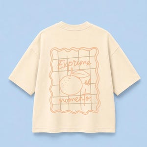 以下が含まれることがあります： 背面にグラフィックがプリントされたクリーム色の半袖Tシャツ。波打つ縁取りの四角いフレームの中に、格子模様、オレンジのイラスト、そして「Exprime el momento」という文字が淡いオレンジ色で描かれています。