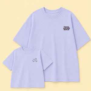 Könnte beinhalten: Zwei hellviolette T-Shirts mit kurzen Ärmeln und Rundhalsausschnitt. Das größere Shirt zeigt den schwarzen Schriftzug TRAVEL EXPLORE REPEAT auf der Brust, während das kleinere Shirt eine kleine schwarze Strichzeichnung eines Rollers zeigt.
