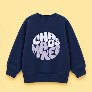 Könnte beinhalten: Ein langärmeliges Sweatshirt in Marineblau mit einer kreisförmigen Grafik in der Mitte der Brust. Die Grafik enthält den Text "CHAOS MAKER" in einer wellenförmigen, fetten, hellvioletten Schrift, die kreisförmig auf dem dunkelblauen Stoff angeordnet ist.