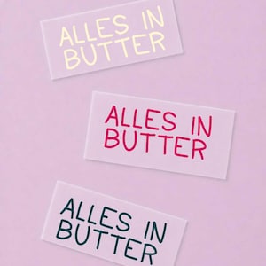Könnte beinhalten: Drei rechteckige Aufkleber mit hellviolettem Hintergrund. Jeder Aufkleber zeigt den Text "ALLES IN BUTTER" in einer anderen Farbe: einer in blassem Gelb, einer in leuchtendem Pink und einer in dunklem Petrol, alle in einer einfachen, runden Schrift.