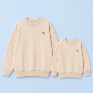 Könnte beinhalten: Zwei beige Rundhals-Sweatshirts mit einem kleinen, dunkelbraun gestickten Croissant-Symbol auf der linken Brust. Beide Kleidungsstücke haben lange Ärmel mit gerippten Bündchen und einen passenden gerippten Saum und sind nebeneinander abgebildet.