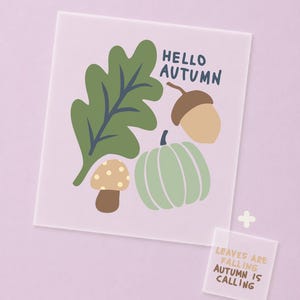 Könnte beinhalten: Ein quadratisches, durchscheinendes Schild mit einem grünen Eichenblatt, einer braunen Eichel, einem beigen Pilz mit weißen Punkten und einem hellgrünen Kürbis mit dem Text HELLO AUTUMN. Ein kleineres Schild zeigt den Text LEAVES ARE FALLING AUTUMN IS CALLING.