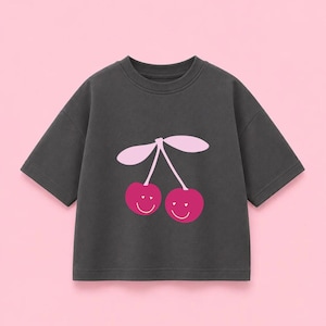 Può includere: Una t-shirt a maniche corte color grigio antracite con una stampa grafica di due ciliegie. Le ciliegie sono rosa scuro con volti sorridenti bianchi e occhi a forma di cuore, accompagnate da due foglie rosa chiaro su un gambo centrale.