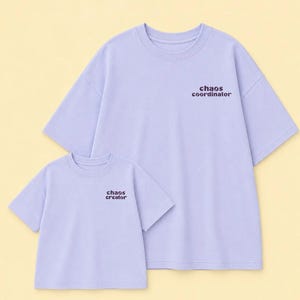 May include: Two light purple t-shirts with short sleeves and crew necklines. The larger shirt features the text "chaos coordinator" in dark brown, while the smaller shirt features the text "chaos creator" in dark brown on the upper left chest.